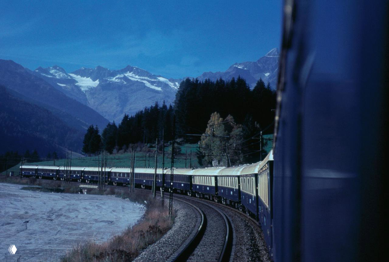 Поезд Venice Simplon‑Orient‑Express в движении по горному маршруту — длинный состав, изгиб рельсов и панорама альпийских склонов на заднем плане.