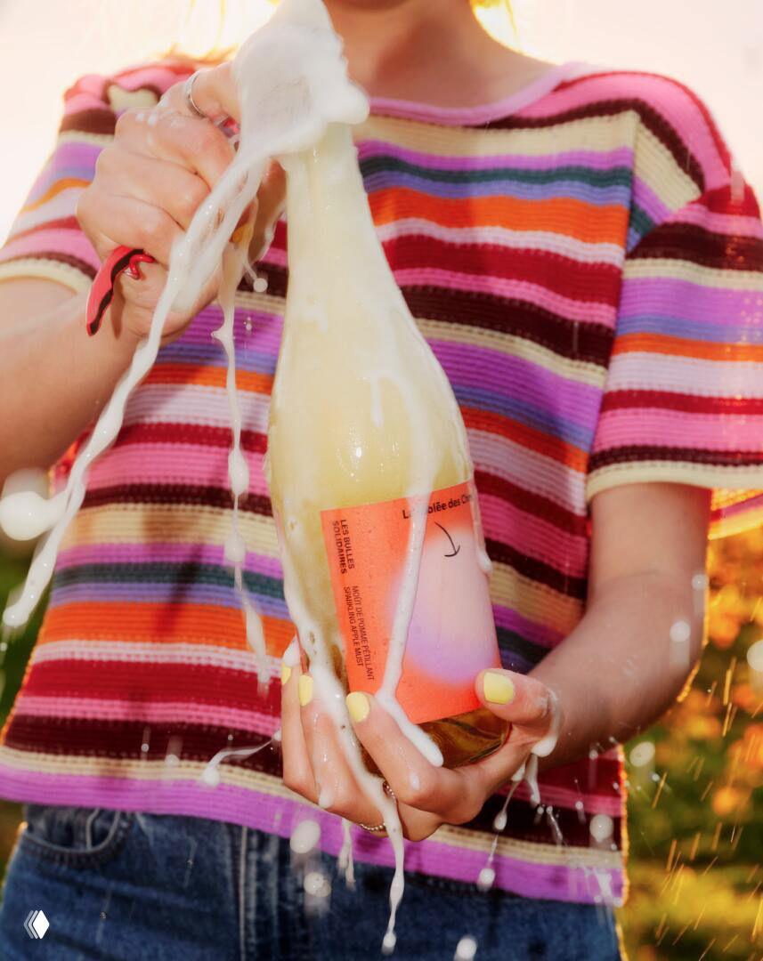 Человек держит вспенившуюся бутылку сидра с яркой пастельной этикеткой на фоне вязаной полосатой футболки, динамичный кадр открытия бутылки.