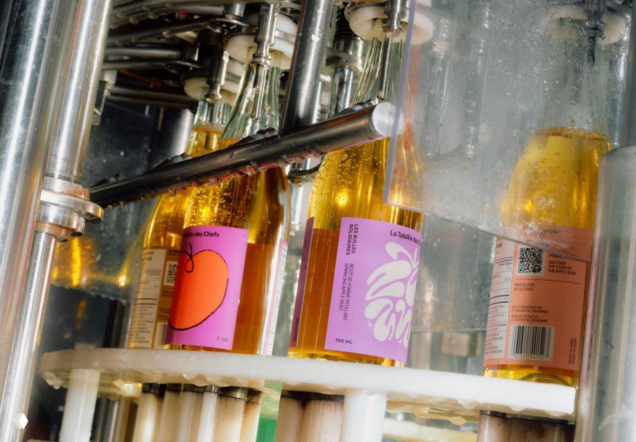 Производственная сцена — бутылки сидра проходят фасовочную линию, видны этикетки с различными иллюстрациями и части оборудования.