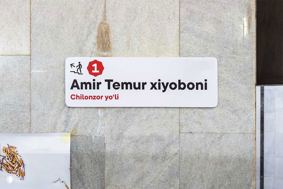 Крупный план настенной таблички «Amir Temur xiyoboni» на светлой каменной облицовке, минималистичный шрифт и пометка линии метро.