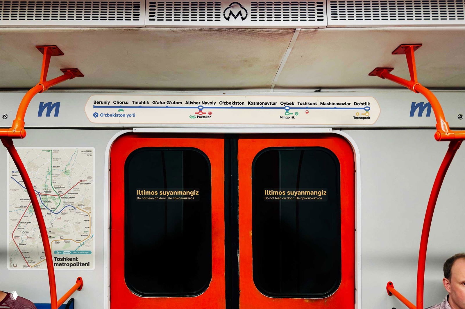 Интерьер вагона с красной рамой дверей, схемой линии над дверями и надписью на узбекском «Iltimos suyanmangiz» — указание безопасности и карта маршрута.