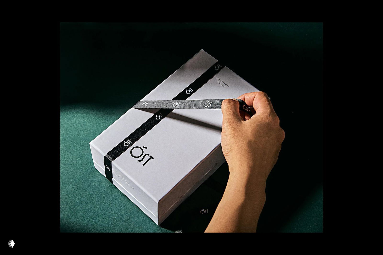Рука завязывает чёрную ленту на белой подарочной коробке с логотипом ÓST — детальный кадр брендирования и материальной упаковки.