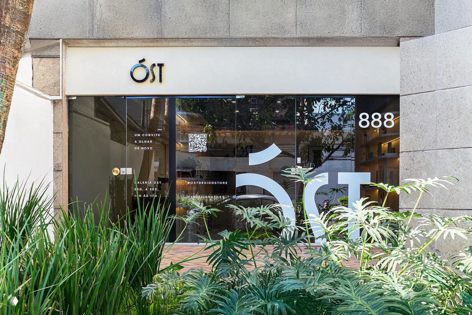 Фасад магазина OST с логотипом на вывеске и крупной стилизованной буквой на стеклянной витрине; зелёная растительность на переднем плане.