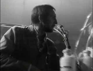 Чёрно‑белый кадр концертного выступления из архива MTV: музыкант у микрофона, характерная плёночная текстура старого видео и атмосферный ракурс.