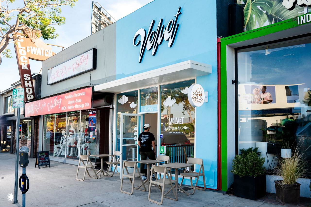 Фасад магазина Velvet в Лос‑Анджелесе с голубой вывеской и скриптовым логотипом; уличная сцена показывает интеграцию айдентики в среду заведения.