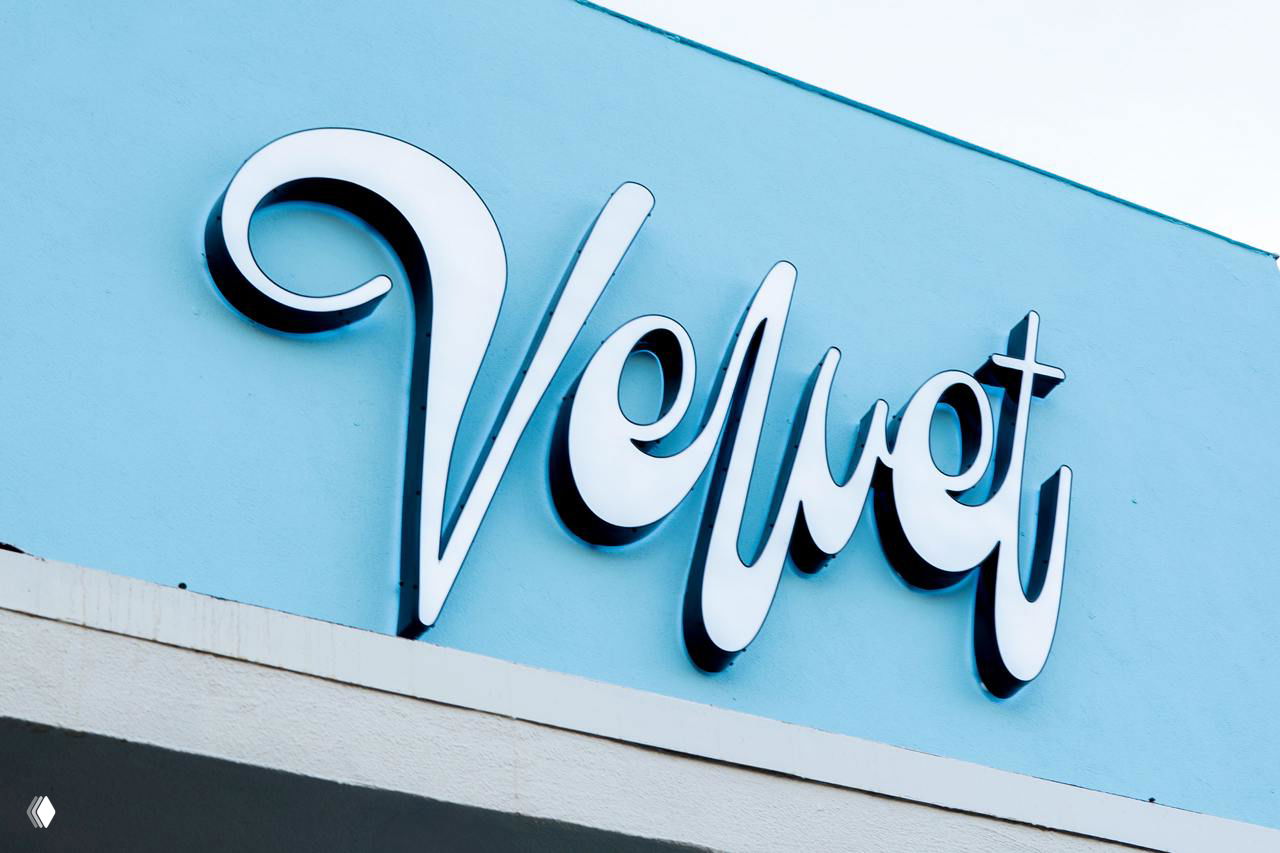 Объёмная металлическая вывеска Velvet на голубом фасаде магазина, игра света и тени подчёркивает скриптовую форму логотипа и наружную айдентику.