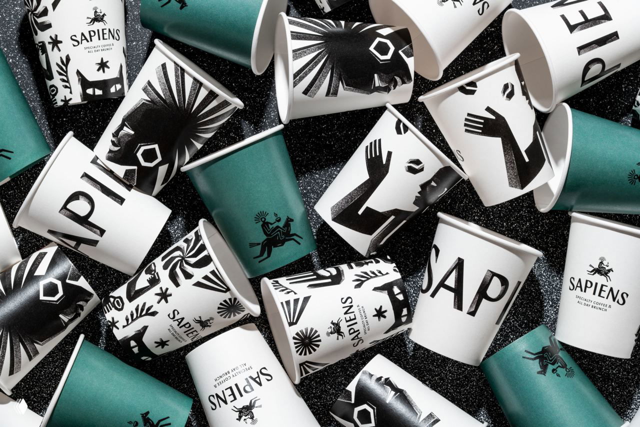 Разбросанные бумажные стаканчики с принтами линогравюр, логотипом Sapiens и зелёными акцентами — композиция упаковки на чёрном фоне.