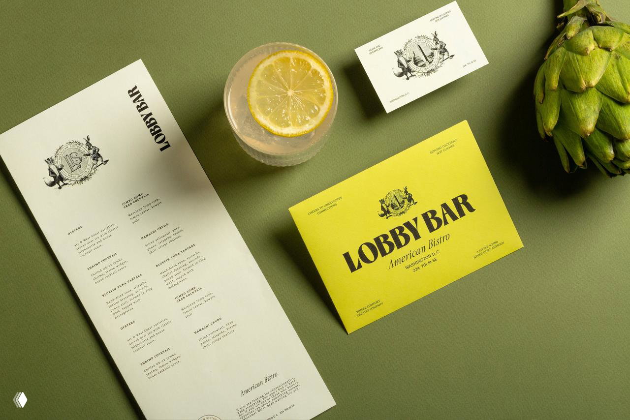Стол с меню, карточками и коктейлем: айдентика Lobby Bar в кадре с лимонными акцентами и золотым тиснением на зелёном переплёте меню.