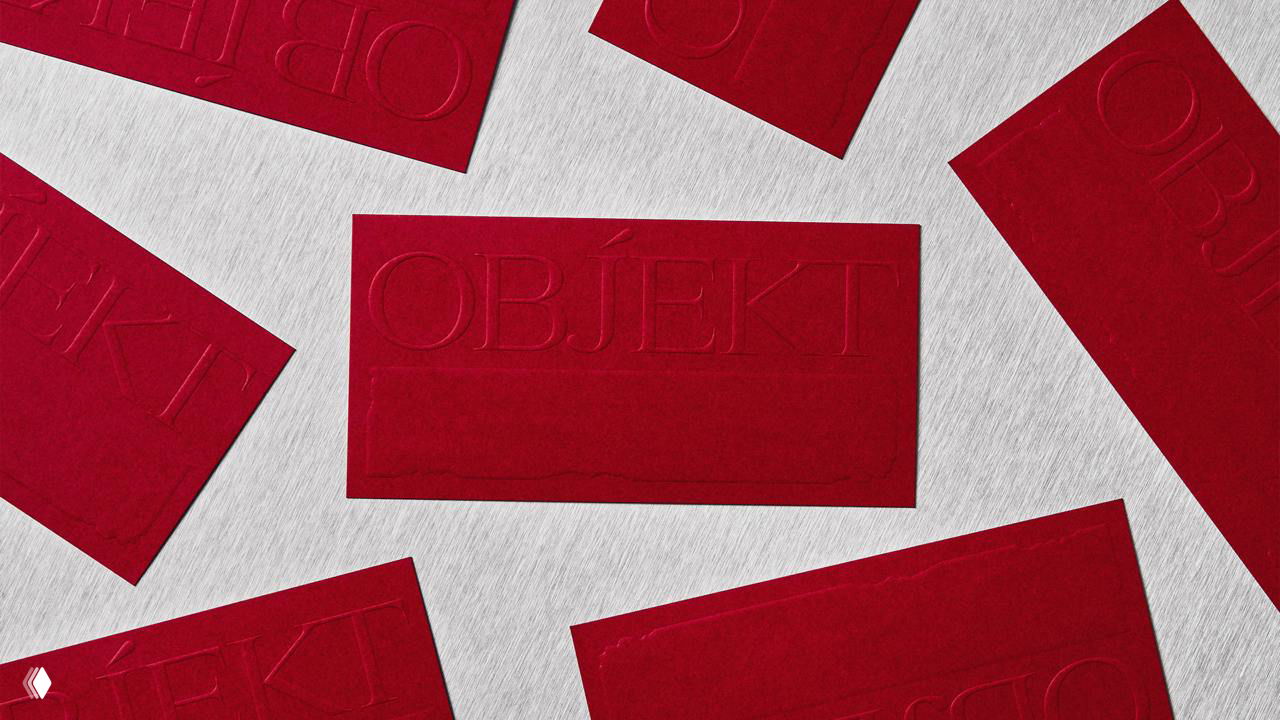 Несколько бордовых карточек OBJEKT, разложенные на светлой текстурной поверхности; видно рельефное тиснение и плотную фактуру бумаги.
