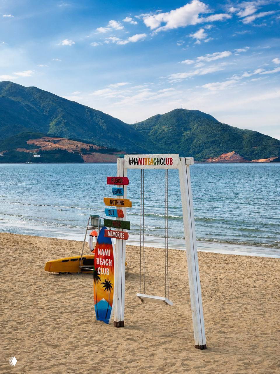 Пустынный песчаный пляж с качелями и яркой табличкой NAMI BEACH CLUB, спокойное море и горы на заднем плане, фотозона на берегу.