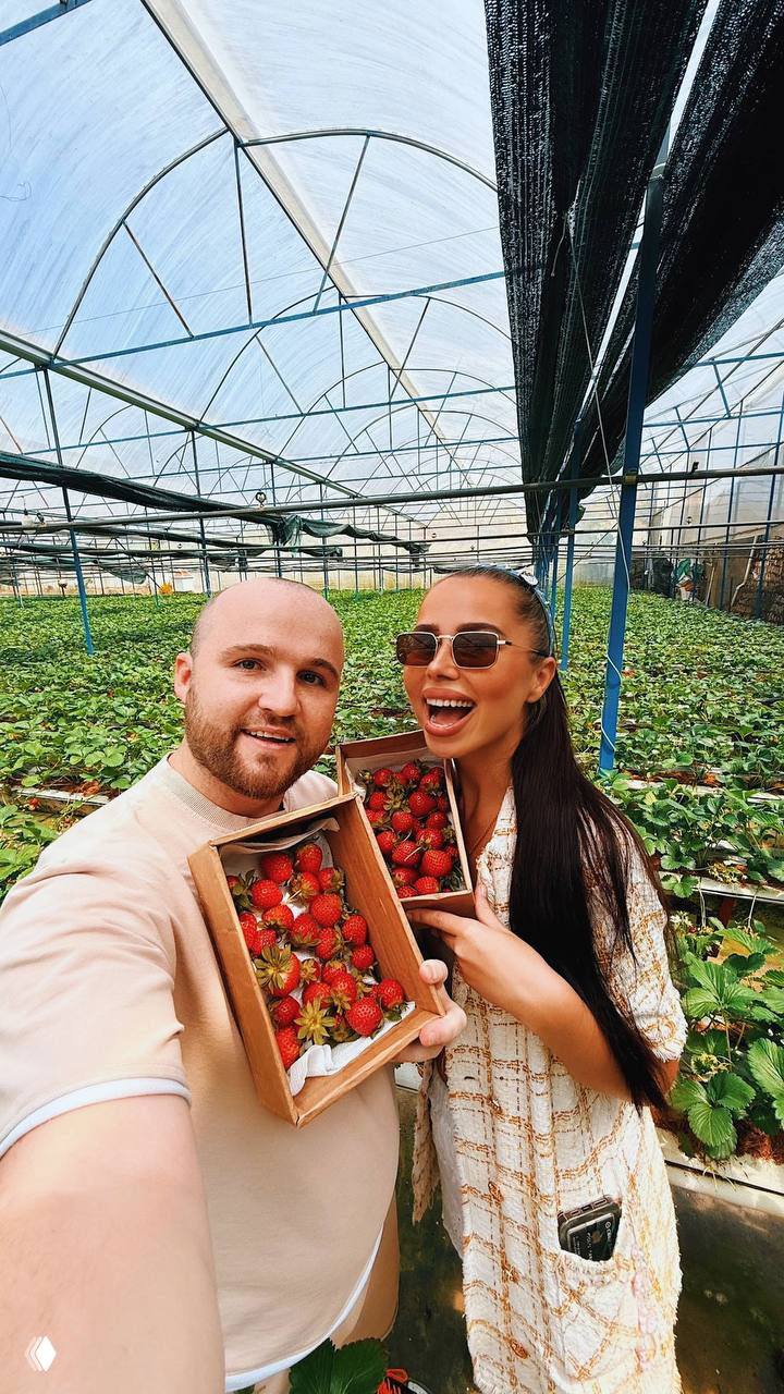 Парное селфи: женщина и мужчина держат коробки с клубникой в теплице, оба улыбаются на фоне аккуратных рядов растений и арочной конструкции парника.