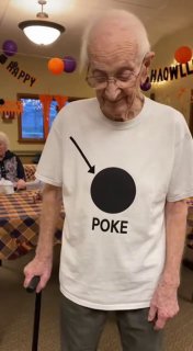 Пожилой мужчина в футболке с надписью «POKE» на праздничном мероприятии: интерьер столовой, украшения и другие участники праздника на заднем плане.