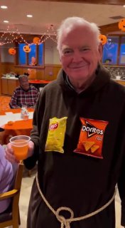 Пожилой мужчина улыбается, на свитере прикреплены пакеты с чипсами Doritos; атмосфера хэллоуин-вечеринки с декором и столами.