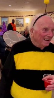 Пожилой мужчина в костюме пчелы держит стакан и улыбается; видна праздничная декорация и другие участники мероприятия.