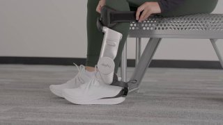 Человек примеряет Dephy Sidekick: лодыжный экзоскелет закреплён на кроссовке, видна посадка устройства и точка крепления на ноге, демонстрация в помещении.