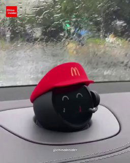 Робот‑компаньон NIO в красной бейсболке с логотипом McDonald’s на приборной панели; вид через лобовое стекло во время дождя.