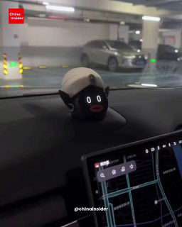 Ночной или подземный паркинг: робот‑фигурка в другой кастомной шляпе на приборной панели, на заднем плане припаркованные автомобили.