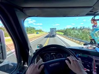 Вид из кабины грузовика: водитель держит руль, видна приборная панель и установленный гоночный сим‑сетап с монитором и педалями.