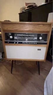 Фронтальный вид ретро-радиолы 1960-х в деревянном корпусе на ножках с панелью управления и решёткой динамика, размещённая в интерьере.