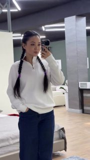 Зеркальное селфи молодой женщины: тёмные волосы в двух косах, белый свитер и джинсы, телефон в руке, видна спальня и зеркало на заднем плане.