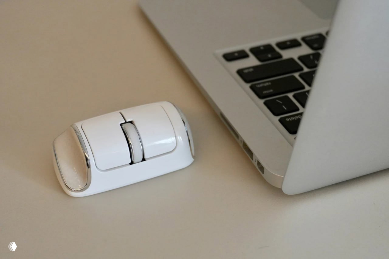 Белая компактная мышь NanoFlow i2 стоит рядом с открытым ноутбуком на столе, подчёркивая её небольшой размер и центральное колесо прокрутки.