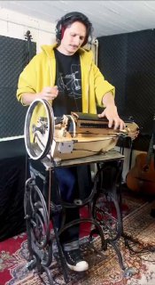 Исполнитель в наушниках ударяет по струнам и регулирует механические элементы швейной лиры; видна педальная станина и ковёр.