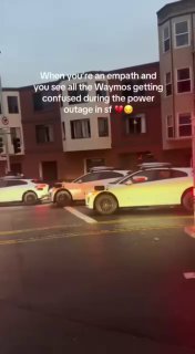 Несколько роботакси Waymo выстроились вдоль тротуара в Сан‑Франциско вечером; подписи на кадре и городские здания на заднем плане при тусклом свете.