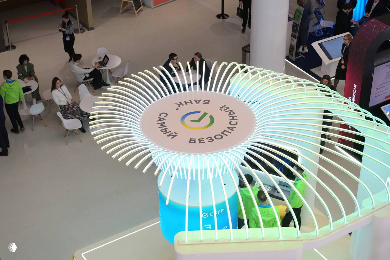 Вид сверху на стенд Сбера с круглой светящейся конструкцией и надписью «Самый безопасный банк», участники форума и зоны общения вокруг, общий план