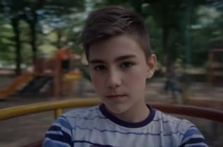 Видео-кадр: мальчик на карусели днём, устойчивое положение тела и выражение лица, движение фона подчёркивает вращение.