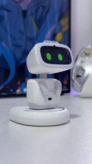 Белый маленький робот‑питомец с тёмным экраном и зелёными «глазами», стоящий на столе на фоне монитора; предметы интерьера размыты.