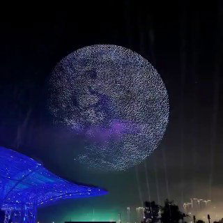 Ночное световое шоу: тысячи дронов в небе над городом формируют большую круглую светящуюся проекцию на фоне городской подсветки и горизонта.