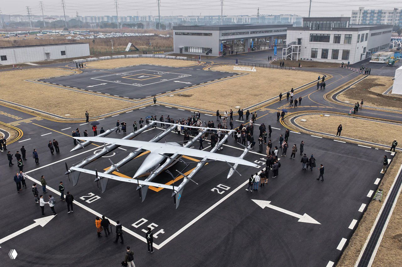 Испытательная площадка AutoFlight: крупный аппарат на земле окружён инженерами и зрителями, видны посадочные площадки и ангар на фоне.