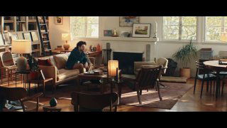 Интерьерный кадр из ролика: мужчина сидит в гостиной за столом, вокруг видна мебель, книги и лампа, создающие уютную бытовую атмосферу.