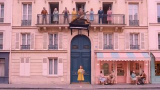 Пастельная улица: женщина в жёлтом платье у входа в розовое кафе, люди на балконах; кинематографичный кадр, иллюстрирует новость о видео.