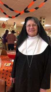 Пожилая женщина в костюме монахини на фоне хэллоуин-декораций в столовой дома престарелых; гости и столы заметны позади.