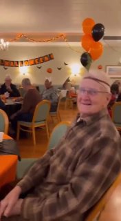 Пожилой мужчина в очках сидит за столом с шариками и праздничными украшениями, атмосфера групповой встречи в доме престарелых.