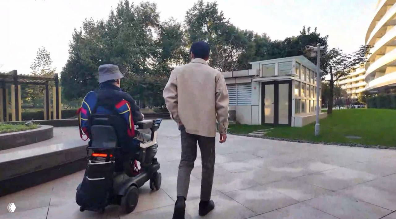 Человек в электрическом инвалидном кресле едет по парковой аллее рядом со сопровождающим, вид сзади, на фоне деревьев и жилого комплекса.