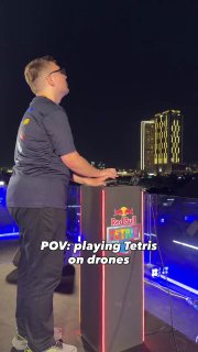 Человек у пульта с логотипом Red Bull на сцене ночью, фон — подсветка и подготовка дронов для игры в огромный Тетрис.