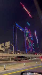 Ночная фотография 150-метровой дубайской рамки, над которой светящиеся дроны формируют фигуры и поле «Тетриса», вид с проезжей части.