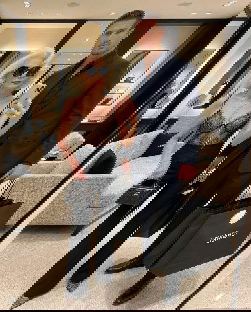 Пенсионерка в стильном образе делает селфи с молодым мужчиной в костюме на фоне бутика; заметны сумки Chanel в руках.