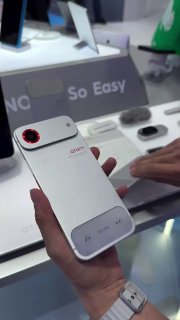 Рука держит белый модульный смартфон Tecno с магнитным модулем на задней панели; на заднем плане видно выставочный стол и аксессуары рядом, демонстрация концепта.