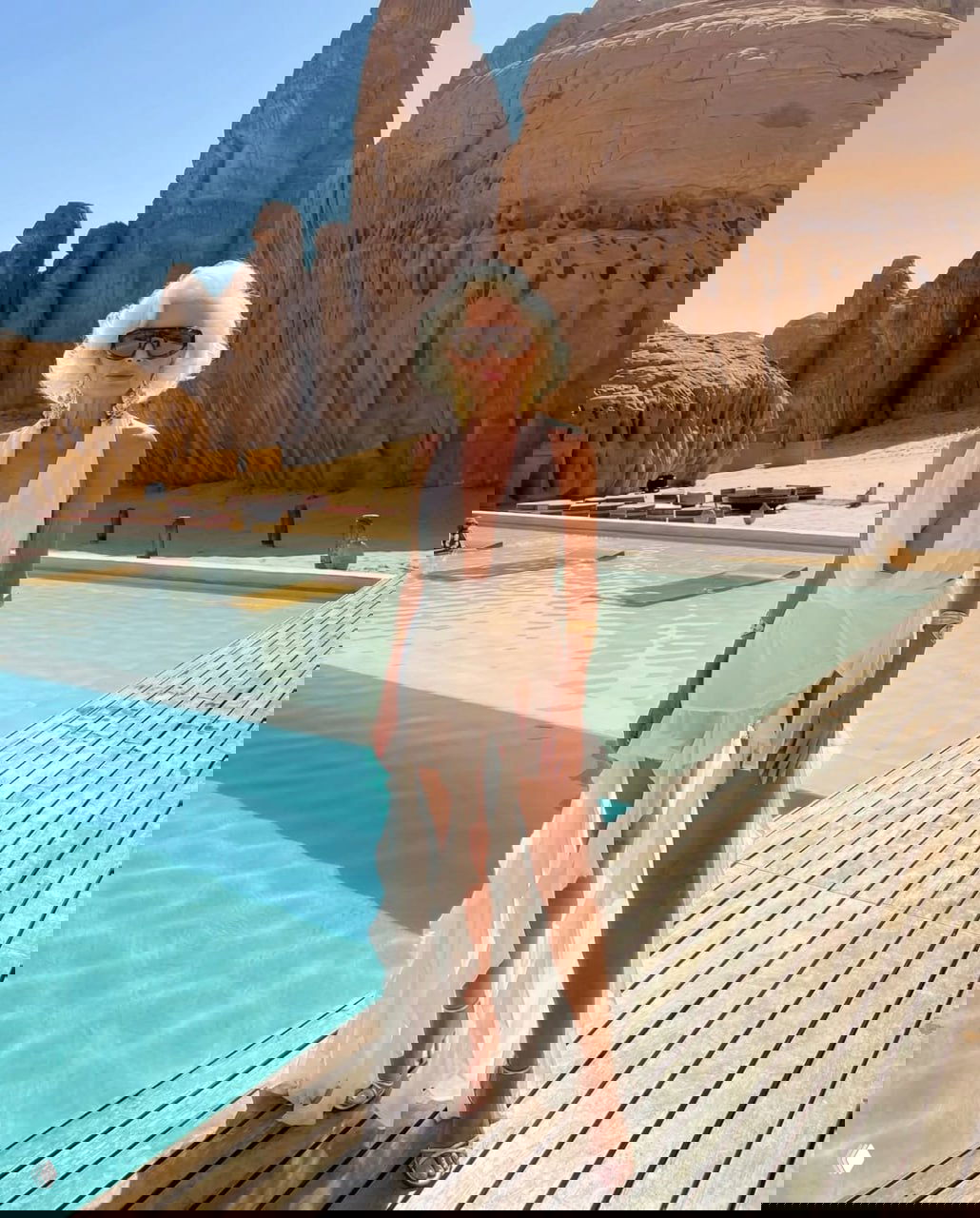 Пенсионерка в белом платье у бассейна на фоне скал и роскошного курорта: пляжный кадр с модным антуражем и солнцем.