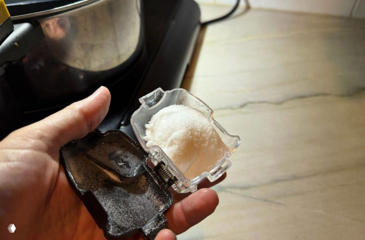 Рука держит прозрачный контейнер с белым порошком (специи/соль) рядом с частью робота, виден механизм дозирования.