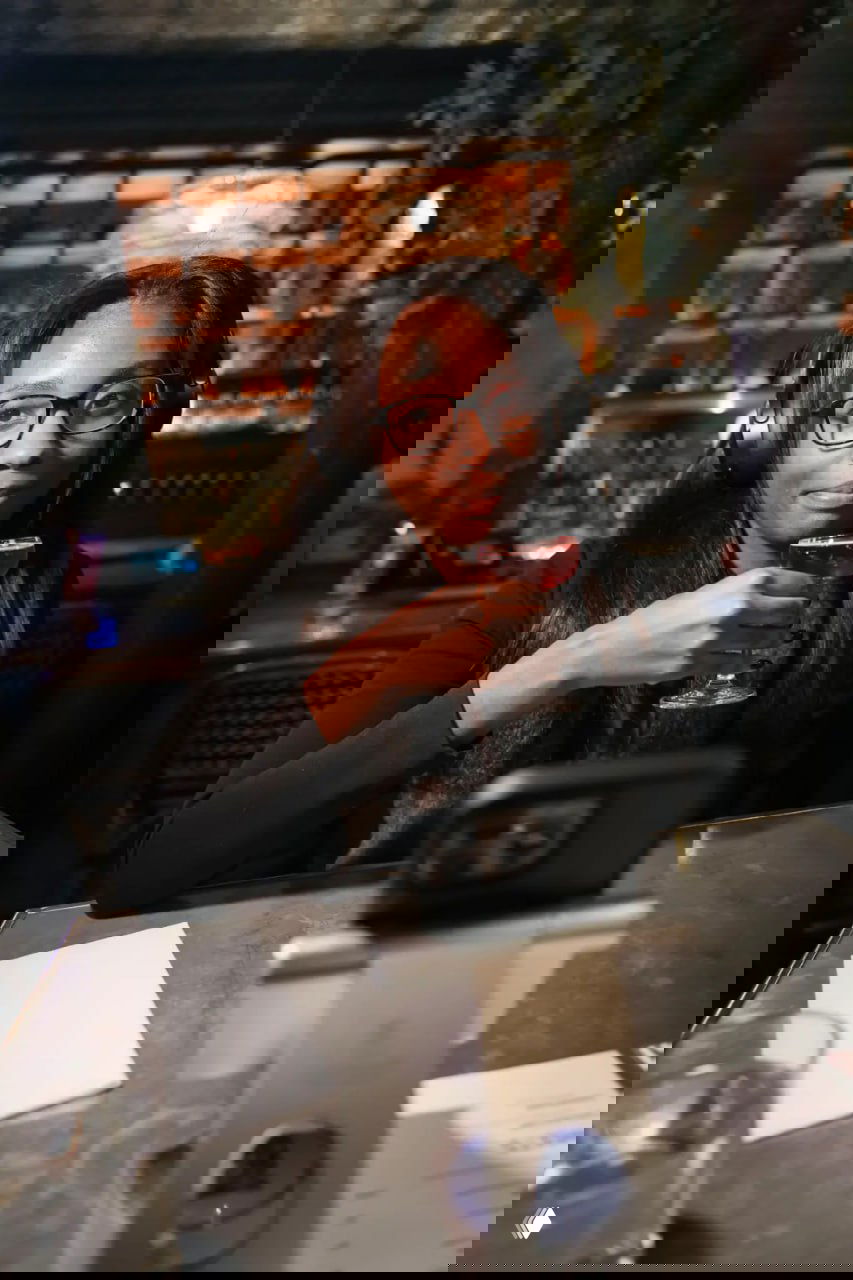 Посетительница в наушниках сидит за столом напротив телефона на подставке и держит коктейль, готовая к видеозвонку с виртуальным партнёром.