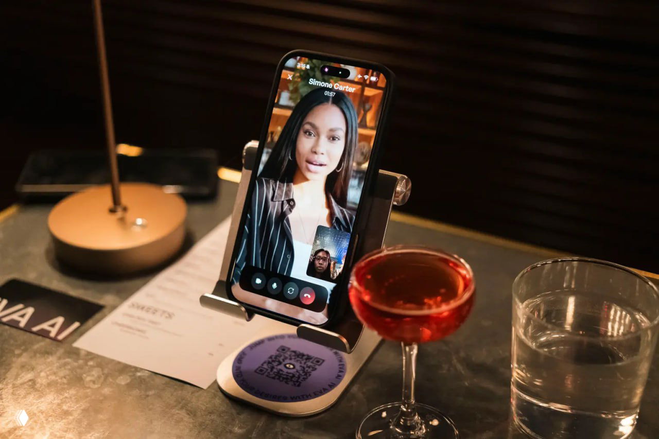 Телефон в подставке показывает видеозвонок с виртуальной собеседницей; на столе бокал коктейля и стакан воды рядом с меню.