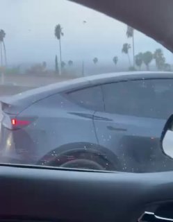 Вид сбоку на серую Tesla на мокрой дороге, снятое из окна другого автомобиля во время дождя; видна задняя часть и бок кузова электрокара.