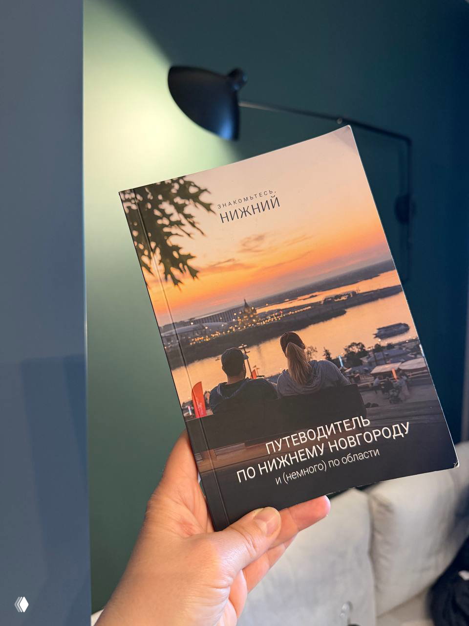 Рука держит путеводитель «Путеводитель по Нижнему Новгороду» на фоне лампы и интерьера номера — обложка с видом на закат и реку.