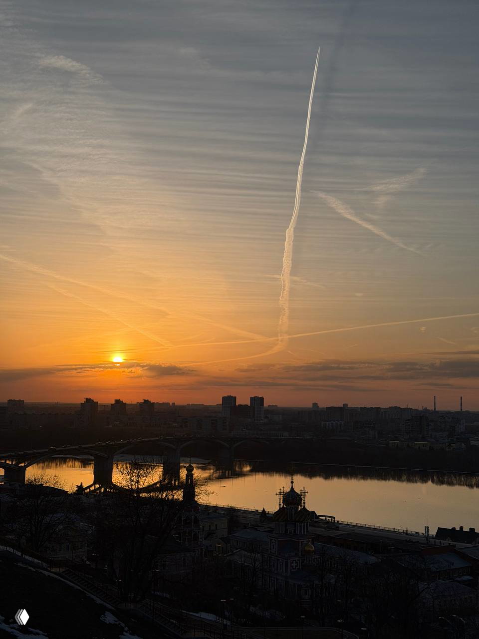 Закат над Нижним Новгородом: солнце на горизонте, силуэты зданий и мост через реку, в небе виден длинный след от самолёта.