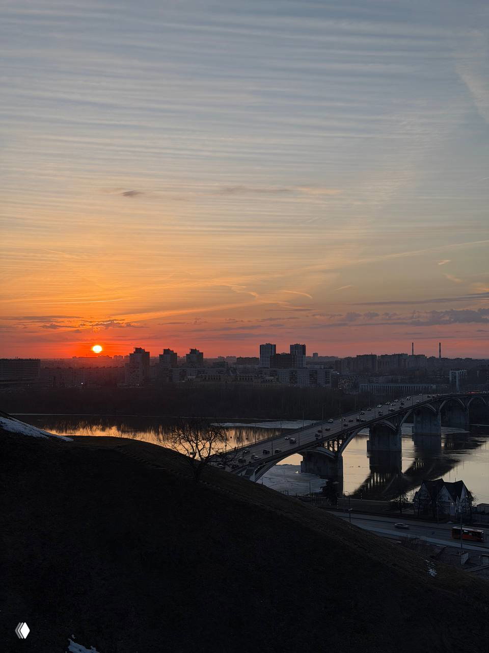 Вид на арочный мост через реку и городской силуэт на фоне тёплого вечернего неба — отражения в спокойной воде и мягкий свет.