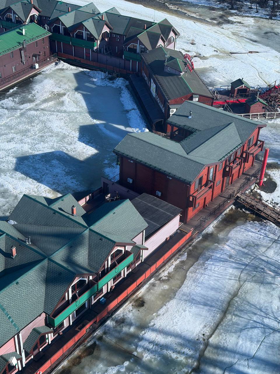 Аэрофотокадр пристани и деревянных зданий у берега реки, весенний лёд и тени домиков — вид сверху со смотровой площадки.